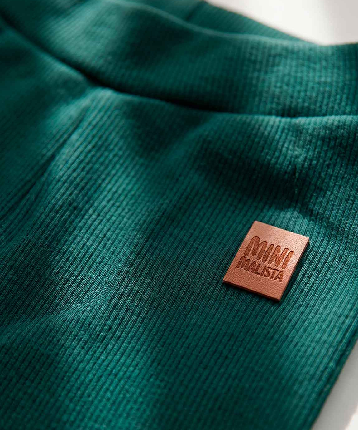 Calça Slim Canelada Bebê Liso Deep Green MiniMalista — cós elástico wide sem cadarço, etiqueta de couro caramelo no hip, malha canelada verde escuro 100% algodão fitted.